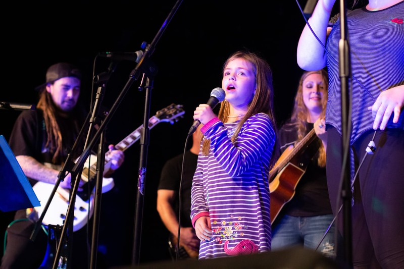 A young girl sings into a microphone while backed up by a band.