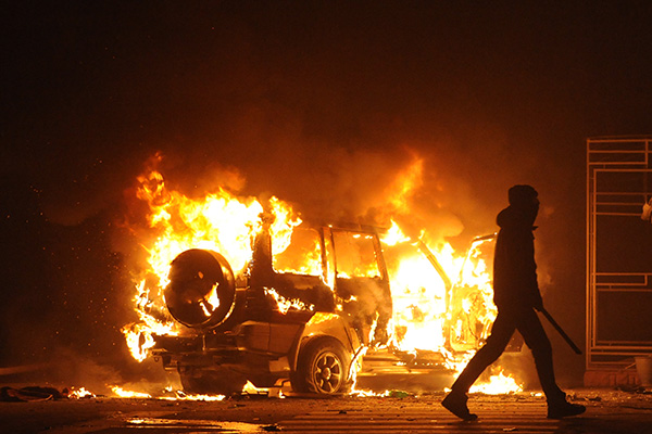 person holding a blunt weapon walks away from a burning vehicle