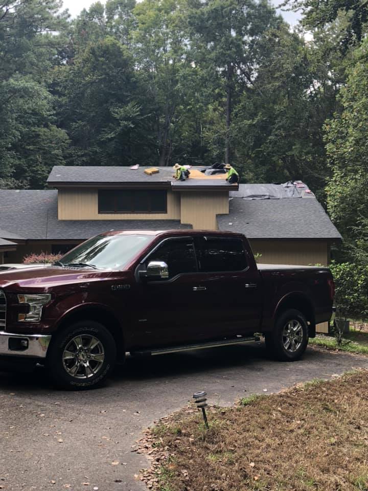 A brown pickup truck sits in front of a house with two workers installing shingles on the upper level of the home’s roof.