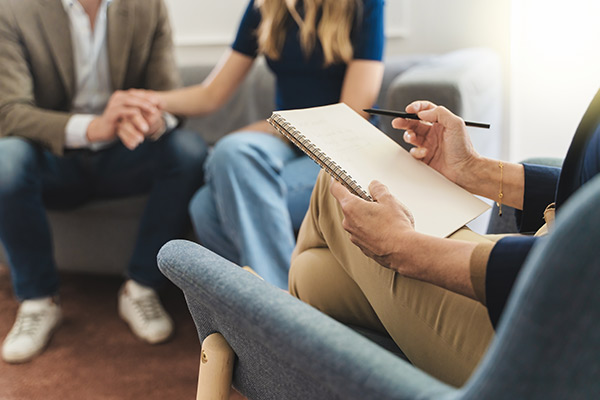 A couple holds hands will talking with a therapist taking notes.