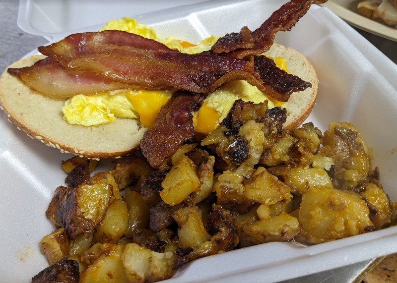 A takeout container of home fries, bacon, and a bagel with eggs.