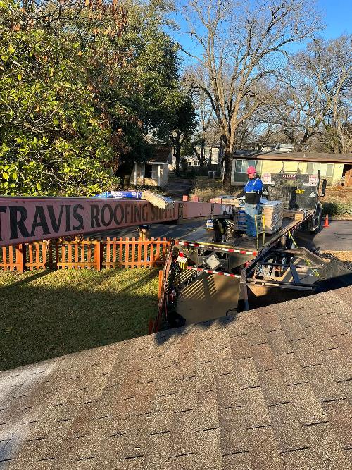 A man stands on a flatbed trailer loaded with piles of shingles and backed up to a house.