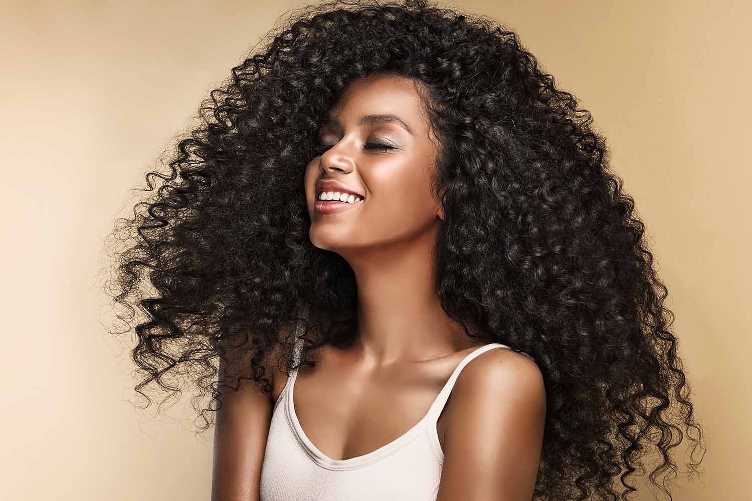 A young black woman with long, curly black hair smiles with eyes closed