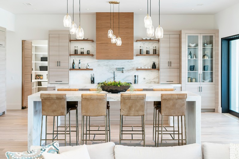 A kitchen scene with four tan chairs pushed up to a white breakfast bar and open cabinetry on the wall.