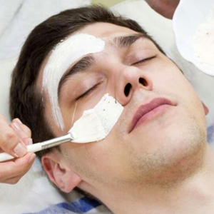 Young man lays with his eyes closed as an esthetician applies facial cream with a brush.