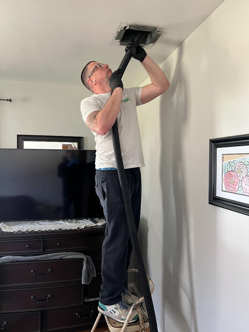 A man stands on a stepladder while reaching a hose into a ceiling air duct.