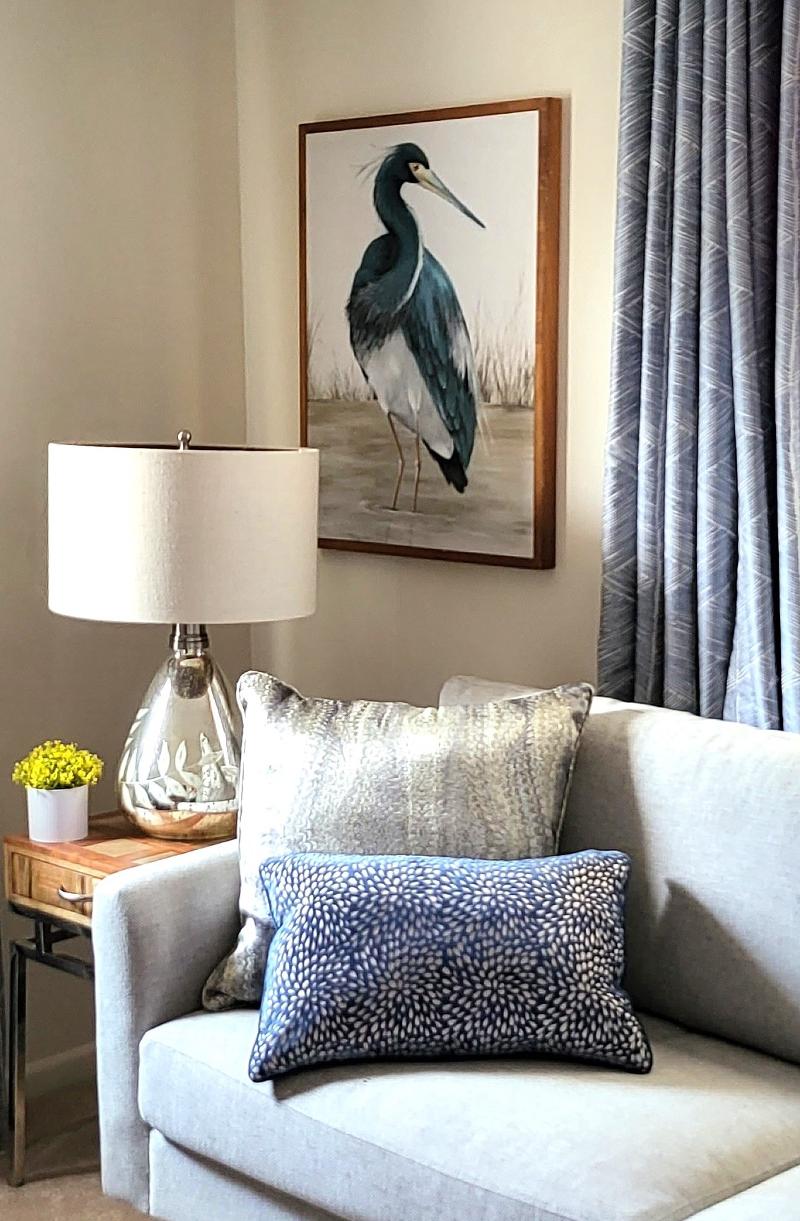 A sitting area with a gray couch, blue curtains, wooden end table, and a painting of a heron.