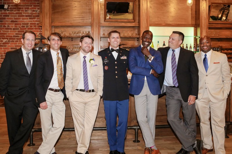 A groom wearing military dress uniform stands with friends and family in formal attire