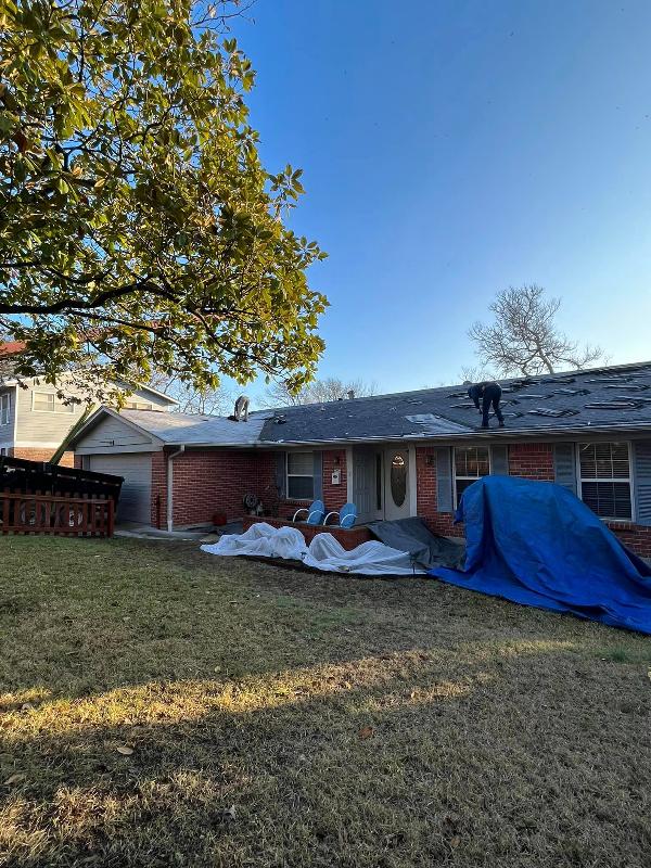 A man performs work on a home’s roof while tarps cover shrubs in front of the house.