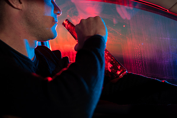 Young man in a car with beer bottle to his lips, illuminated by police lights