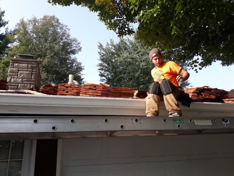 A roofer sits on the edge of a roof while laying roof tile sections.