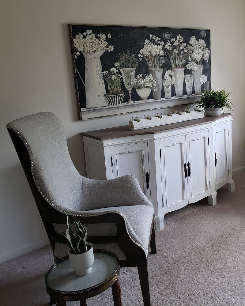 A sitting area with a gray high-back chair, white credenza, and wall painting of flowers.