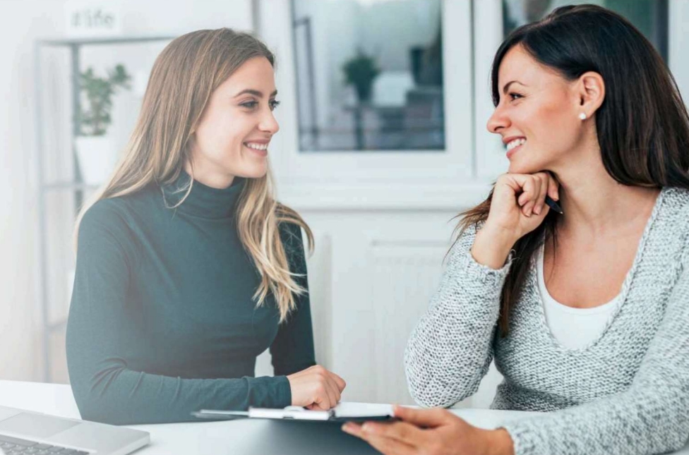 Two women smile at each other as they sit at a table together.