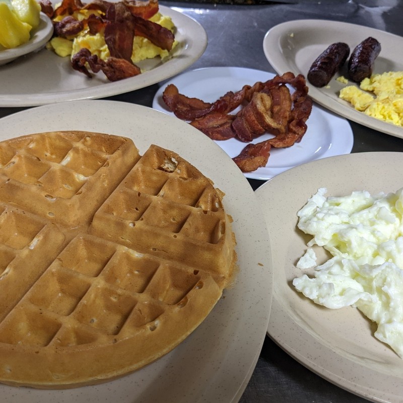 Breakfast plates of a waffle, bacon, sausage, and eggs.