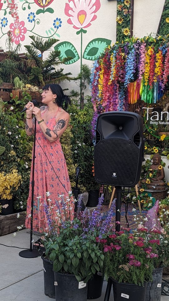 A lady in a long, pink dress speaks into a microphone standing among many green plants.