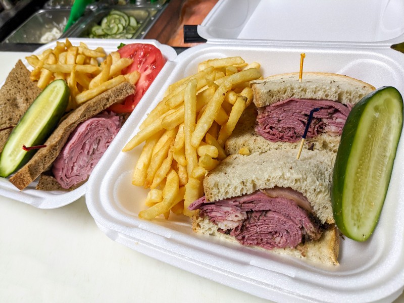 Takeout containers of corned beef sandwiches, fries, and pickles.