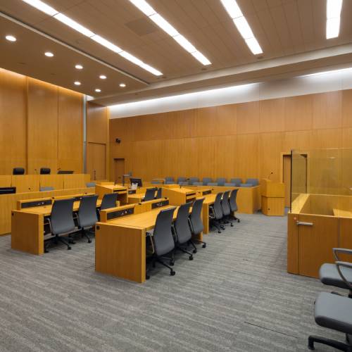 An empty courtroom with tables and chairs, a judge's bench, and gray carpet.