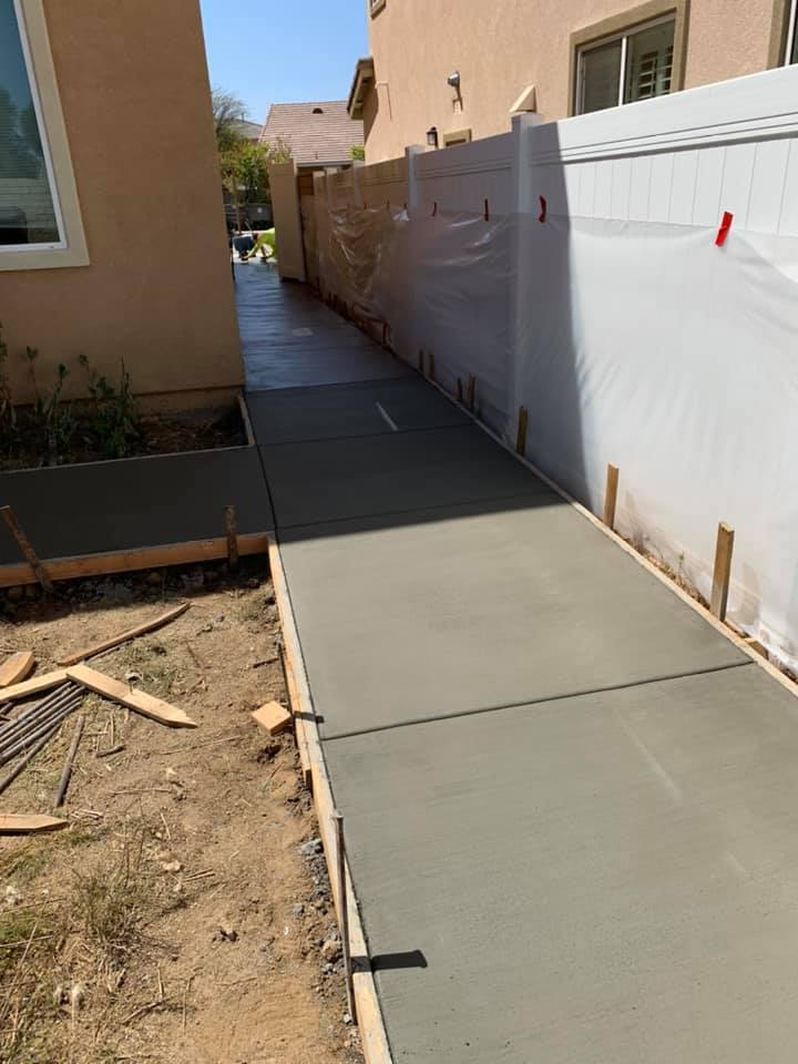 A freshly poured sidewalk running along a white fence and in front of a home.