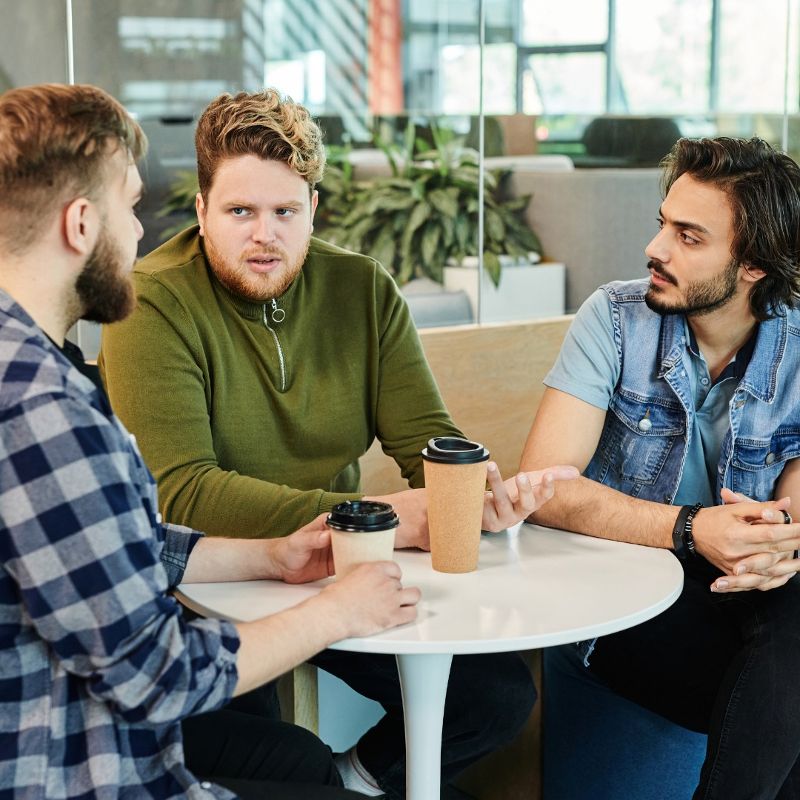 Three men have a discussion while sitting around a small, round table.