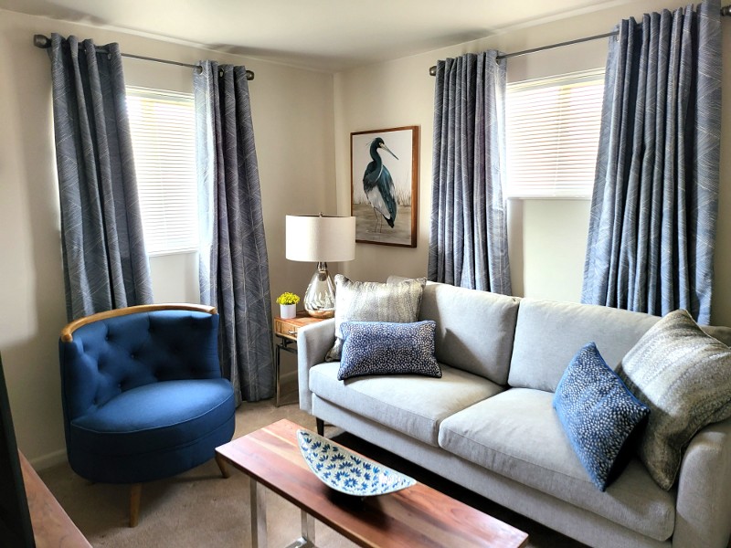 A sitting area with a blue chair, gray couch, blue curtains, wooden end table, and a painting of a heron.