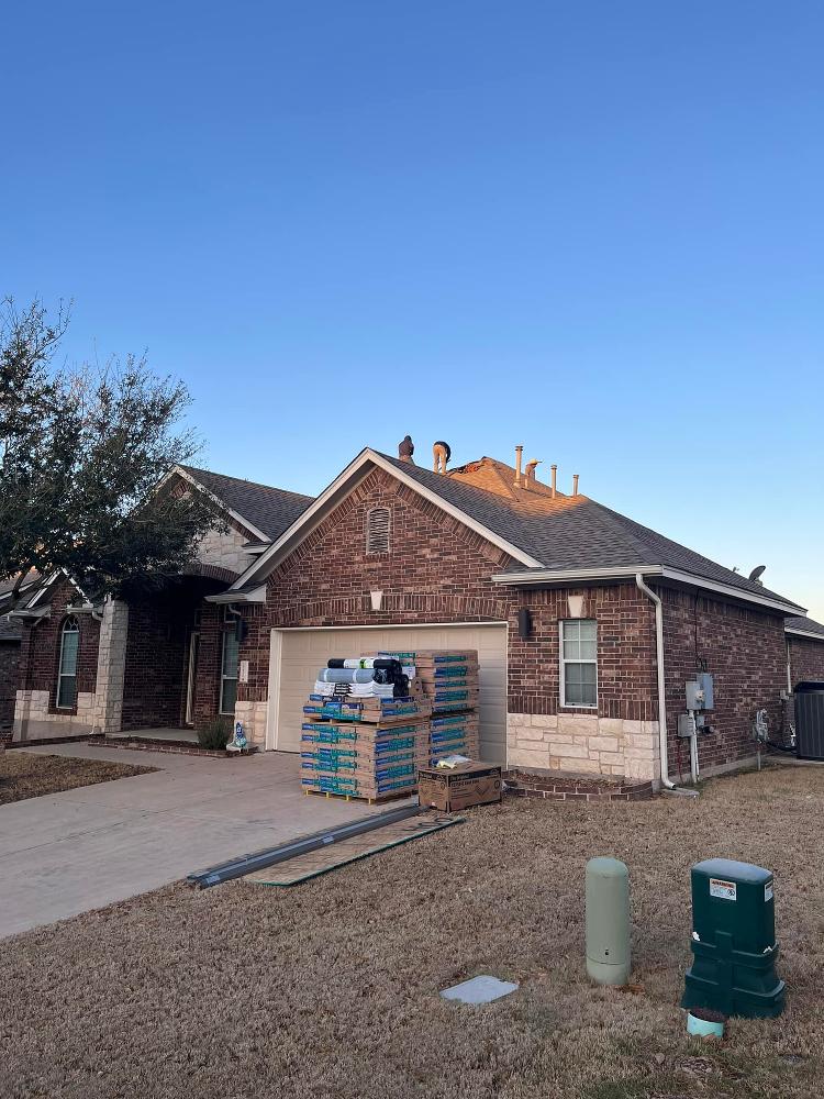 Roofers install a shingle roof on a brick home with stacks of shingles sitting in front of the home’s garage.