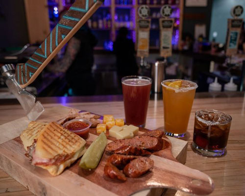 A charcuterie board of a panini, a pickle, sausage, cheese cubes, and dip with three drink glasses.
