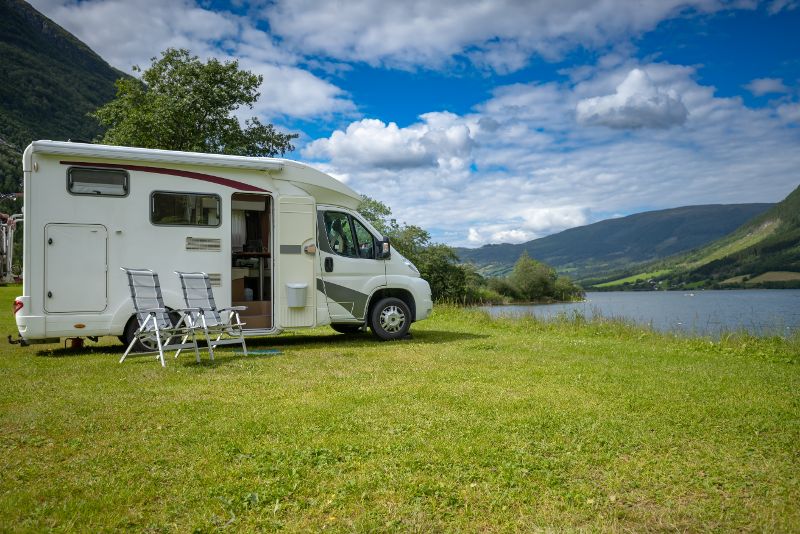 A motor home is parked in a grassy area next to a lake with two folding chairs sitting outside the RV..