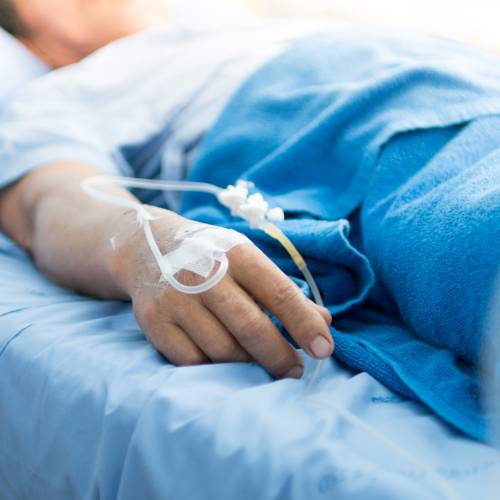 A person lies in a hospital bed, with blue sheet, blue blanket, IV tube