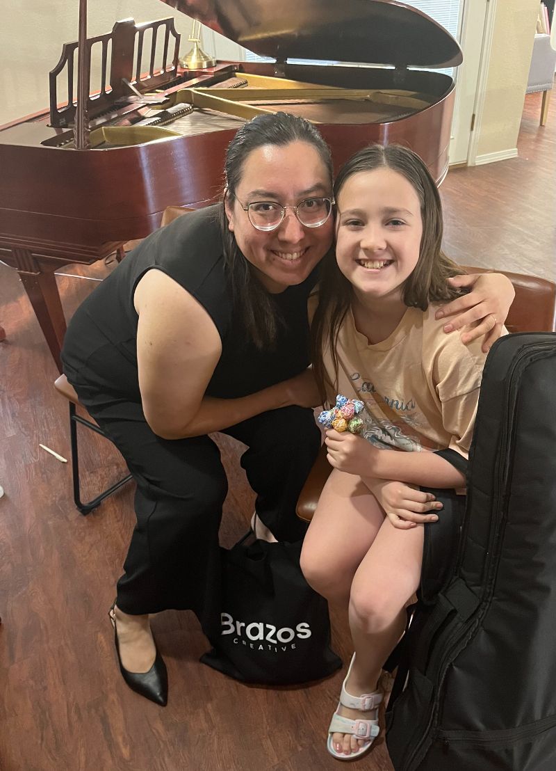 Founder of Brazos Creative, Stephanie, smiling with a student in front of a piano.