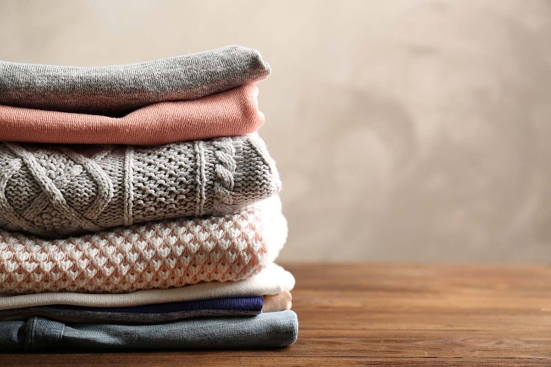 A pile of neatly folded sweaters in neutral tones site atop a wooden table