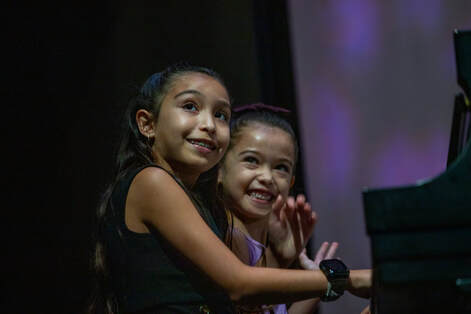 Two young girls smile while sitting at a piano.