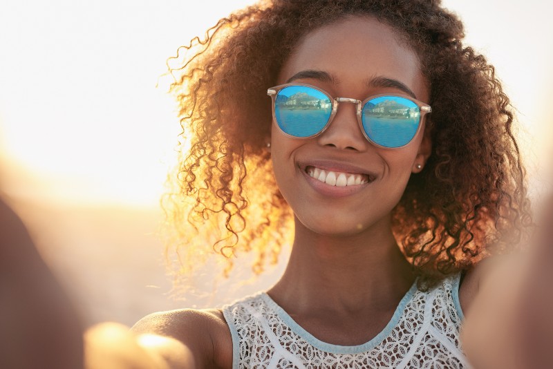A young black woman with curly brown hair and reflective sunglasses smiles at the camera