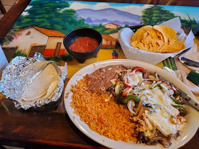 A plate of steak fajitas with peppers covered in a cheese sauce with fried rice and refried beans.