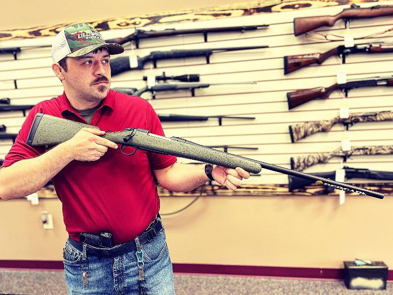 A man holds a shotgun with a wall of shotguns behind him.