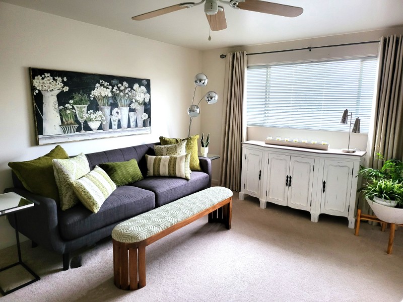 A gray upholstered couch with a kneeling bench and green pillows on the couch.