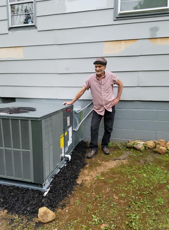 An older man in a brown cap stands next to an outdoor HVAC unit.