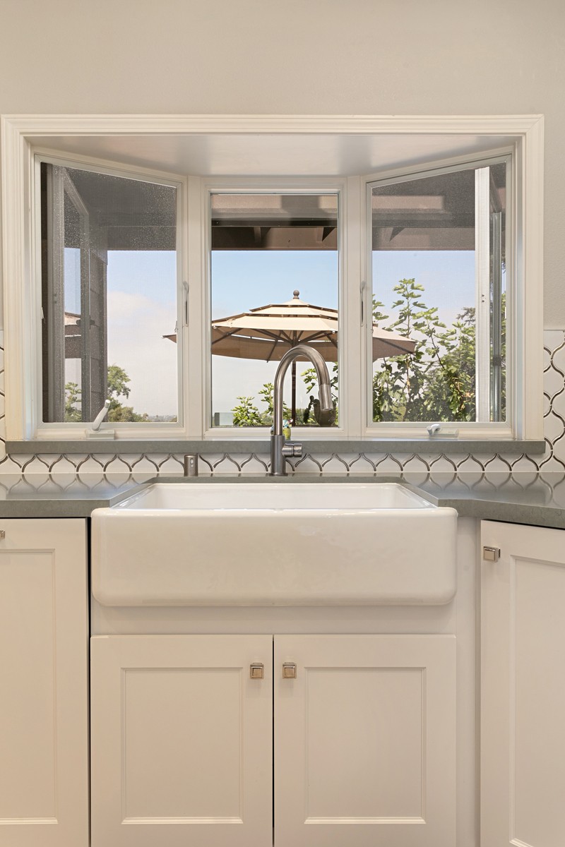 Farm house sink below a small bay window, white cabinets and gray top