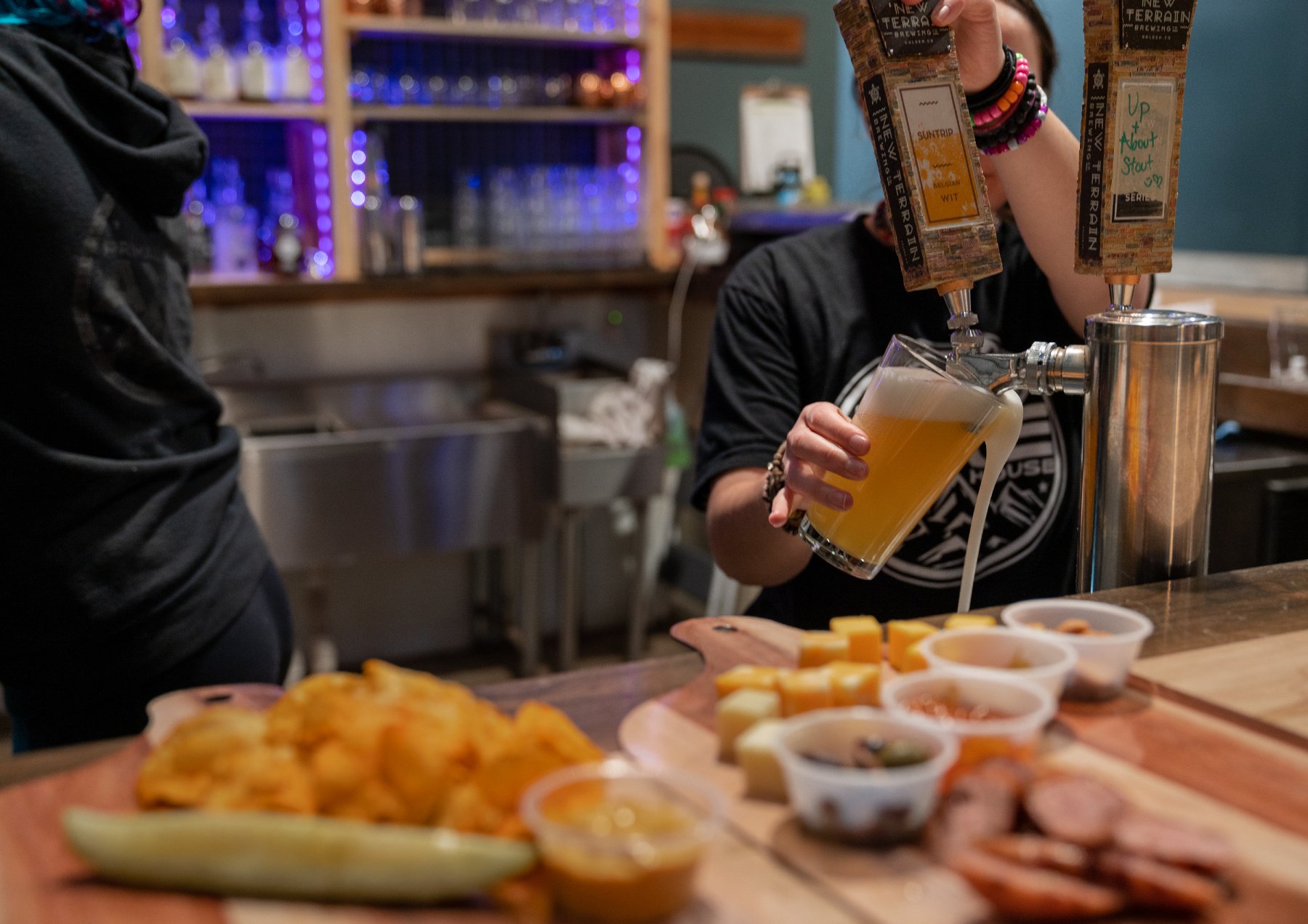 A bartender draws a glass of draft beer with sausage, chips, a pickle, cheese, and dipping sauces in the foreground.