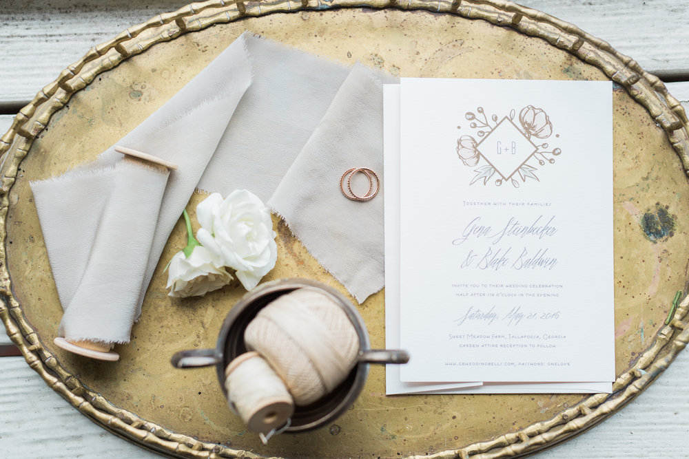 A gold tray holds a wedding invitation and wedding rings lying on a white linen fabric.