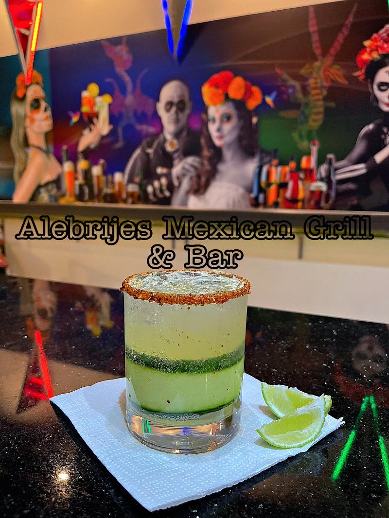 A yellow and green cocktail sits on a table with artwork of Alebrijes Mexican Grill & Bar on the wall.