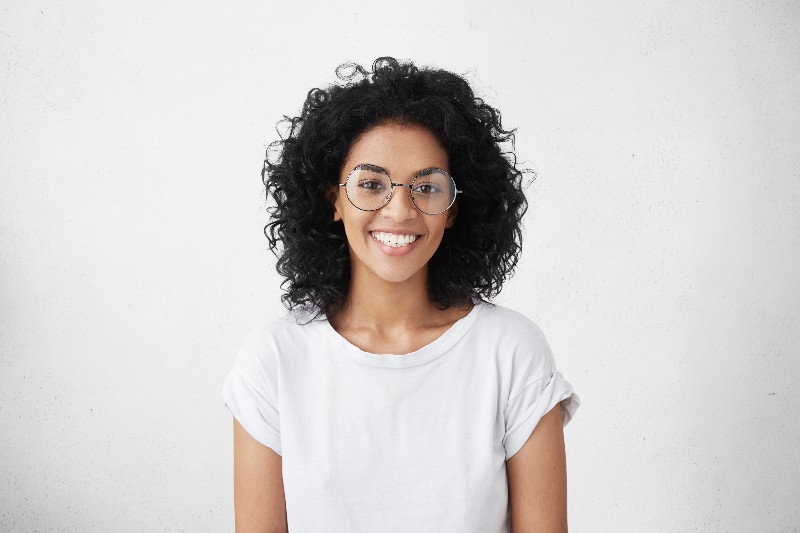 A young black woman with curly brown hair and reflective sunglasses smiles at the camera