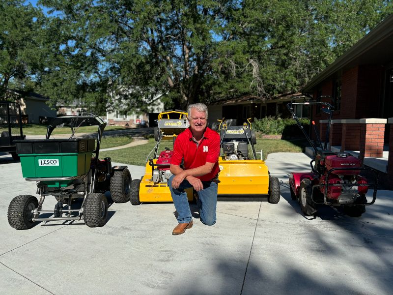 A man kneels on the ground surrounded by lawn and garden equipment.