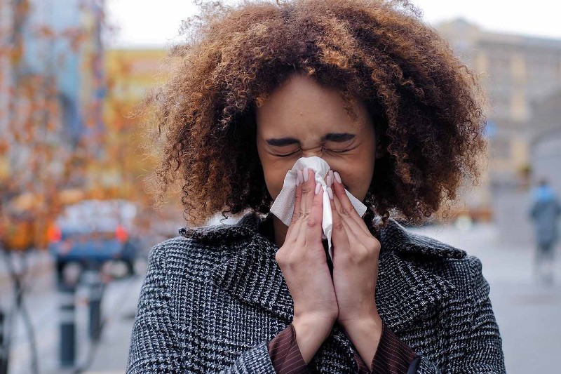 A young woman with curly hair blows her nose while standing on the sidewalk