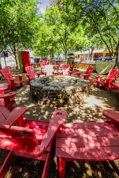 Red Adirondack chairs surround a stone firepit, lush green trees provide shade